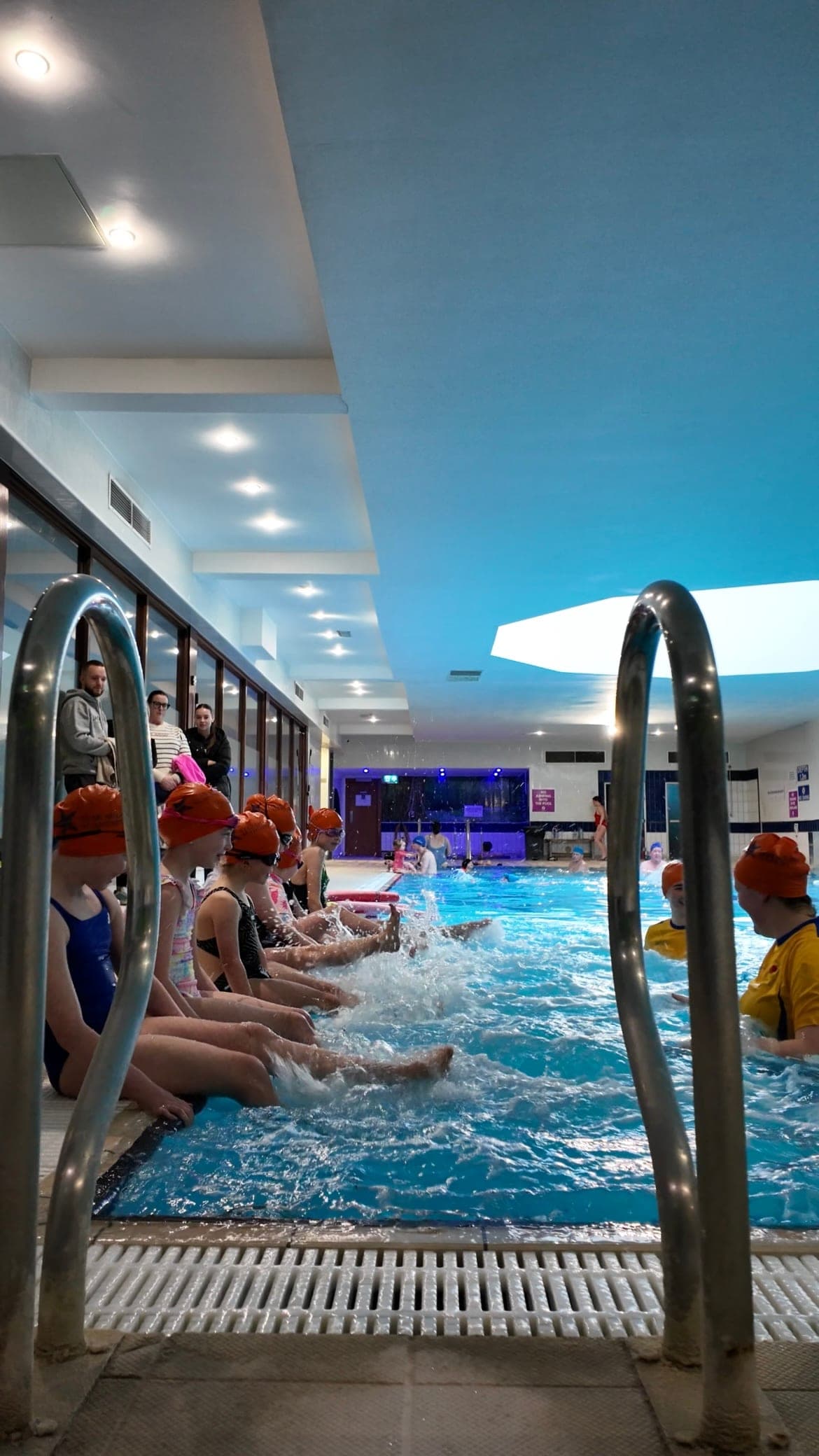A line of swimmers in Star Splash caps sitting on the poolside kicking up spray