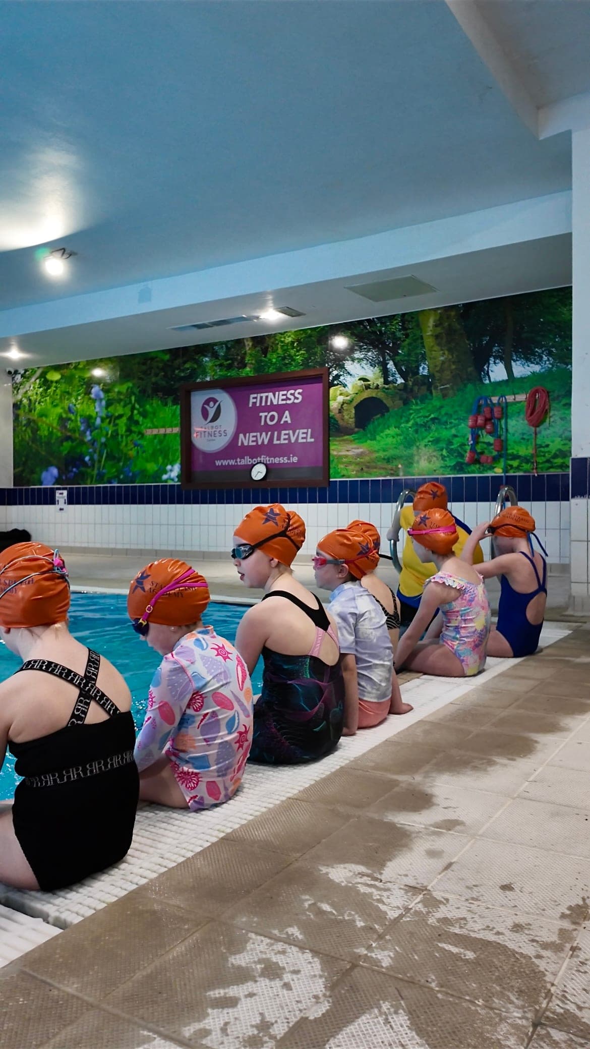 A row of young swimmers in branded orange Star Splash Academy caps along the poolside
