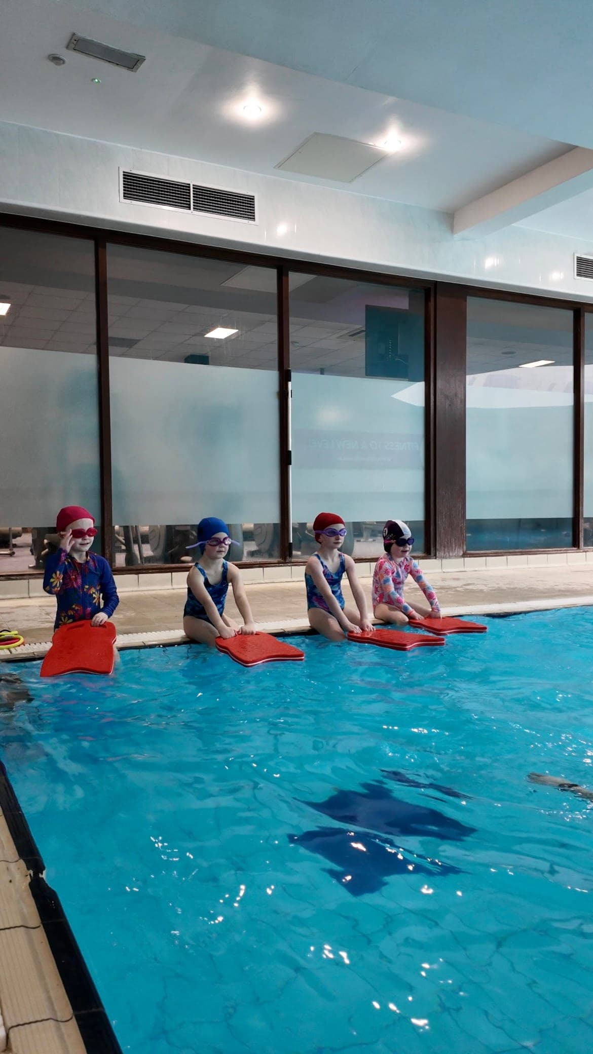 Four swimmers sitting on the edge of the pool holding red kickboards