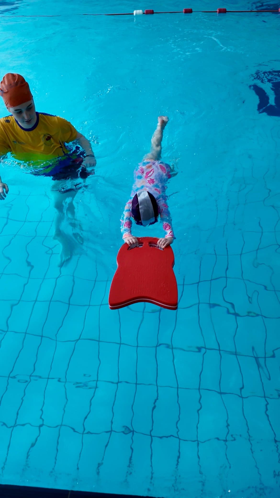 A young swimmer kicking with a red kickboard while their teacher swims alongside